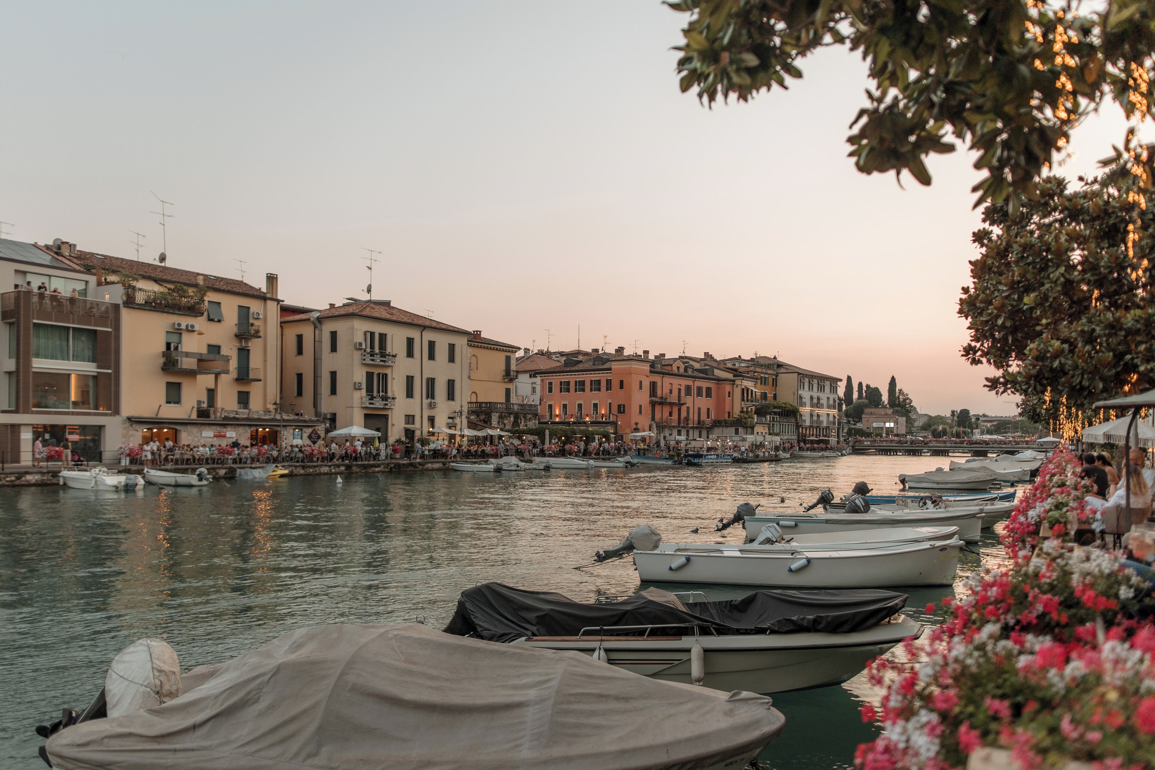 Una passeggiata sul lungomare con barche ormeggiate, edifici con balconi e una folla di persone.