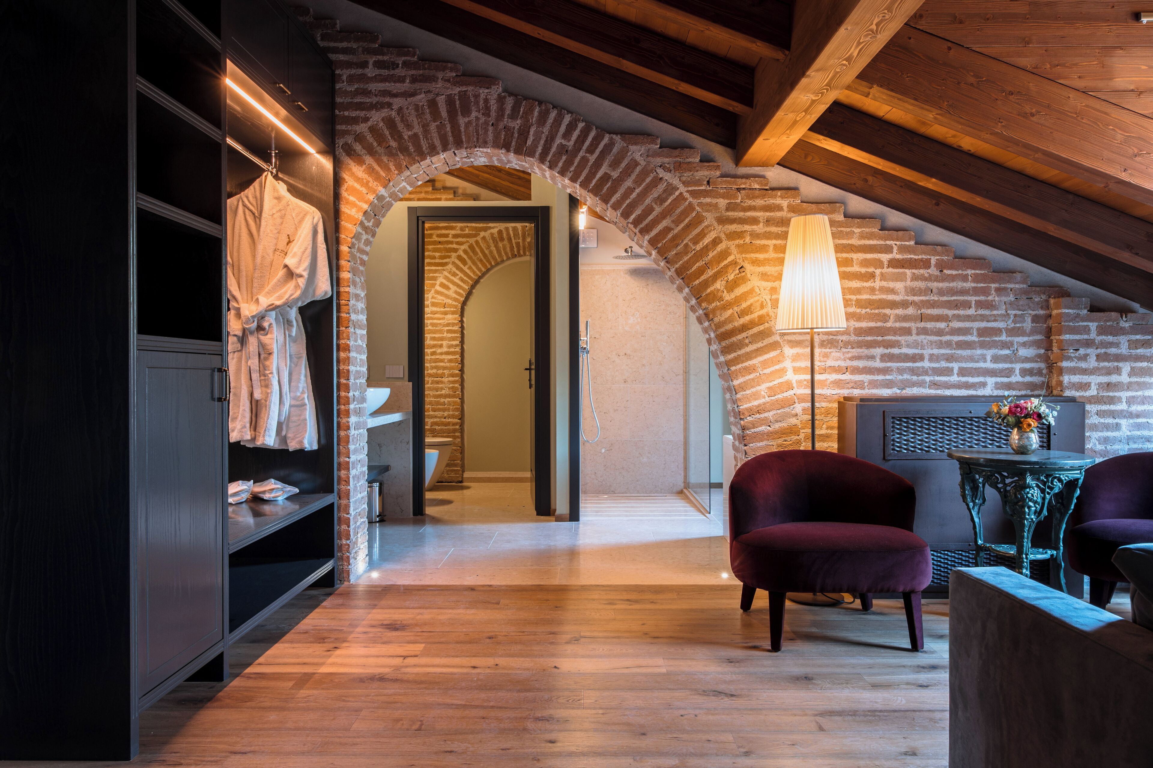 A room with a wooden floor, a brick archway, a hanging robe, a lamp, and two chairs.