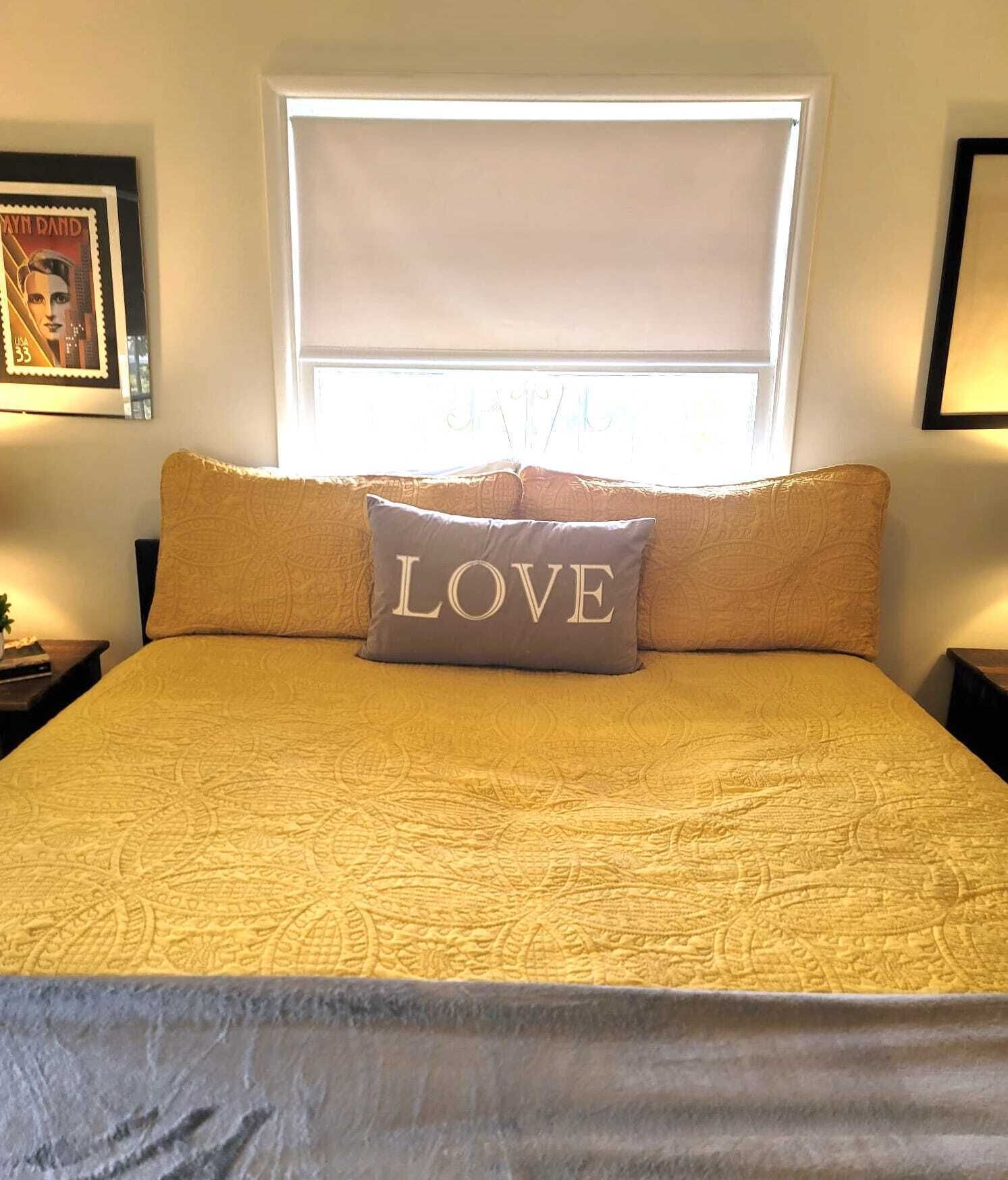 A bed with a patterned bedspread, a pillow with the word "LOVE," and a window with a white blind.