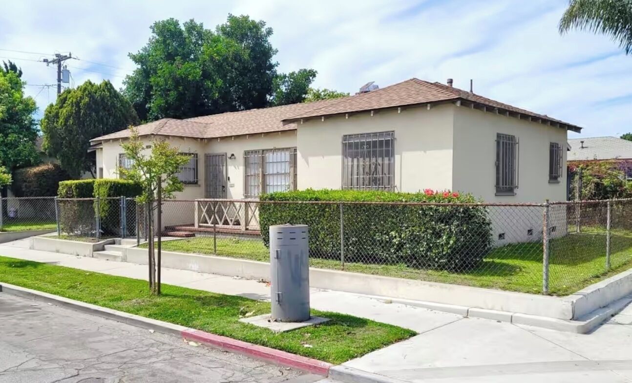 A single-story house with a beige exterior, a tiled roof, and a fenced yard with a small tree and shrubbery.