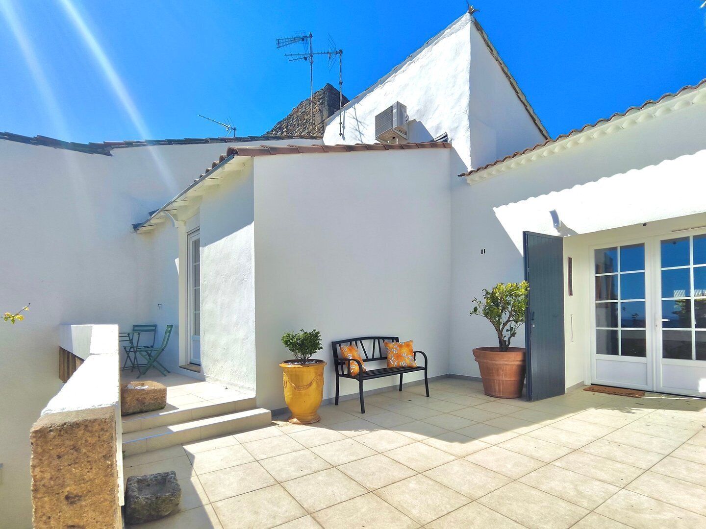 A white building with a tiled patio, a bench, a potted plant, and a door.