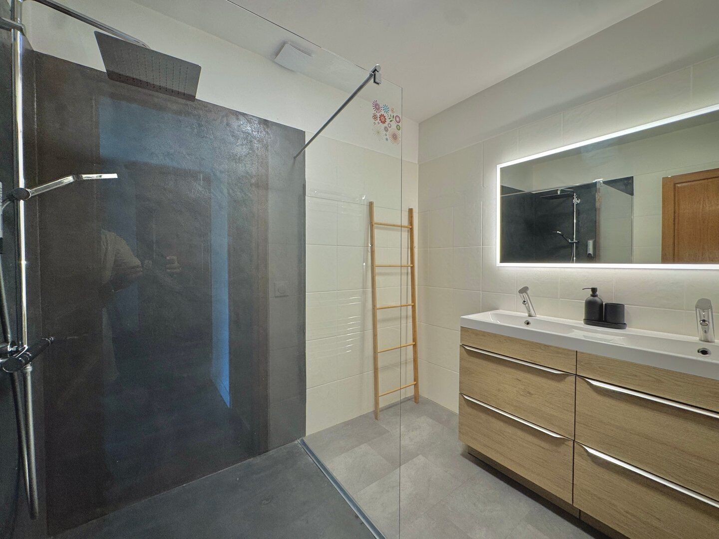 A modern bathroom with a shower, a wooden ladder, a large mirror, and a vanity with drawers.