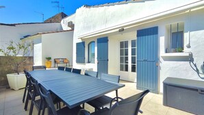 A patio with a table and chairs, a small garden, and a white building with blue shutters.