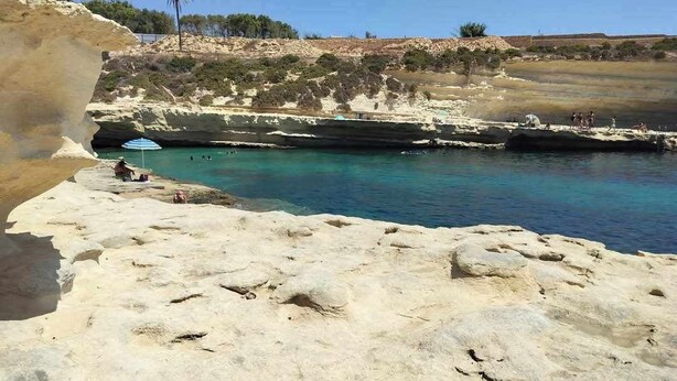 A rocky beach with clear blue water, a person sitting under an umbrella, and a cliff in the background.