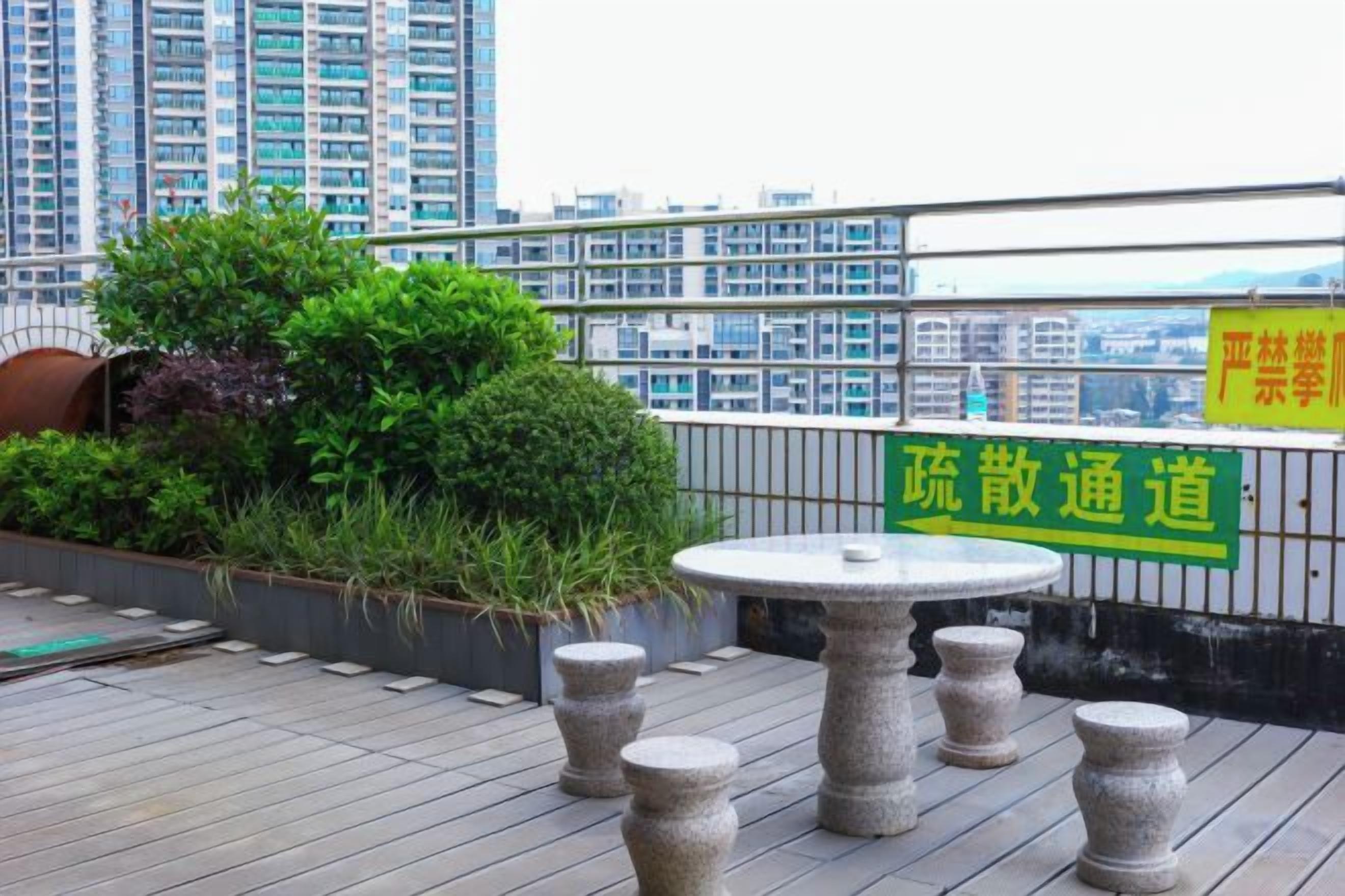 Una terraza en la azotea con una mesa redonda y taburetes, rodeada de vegetación y vistas a rascacielos.