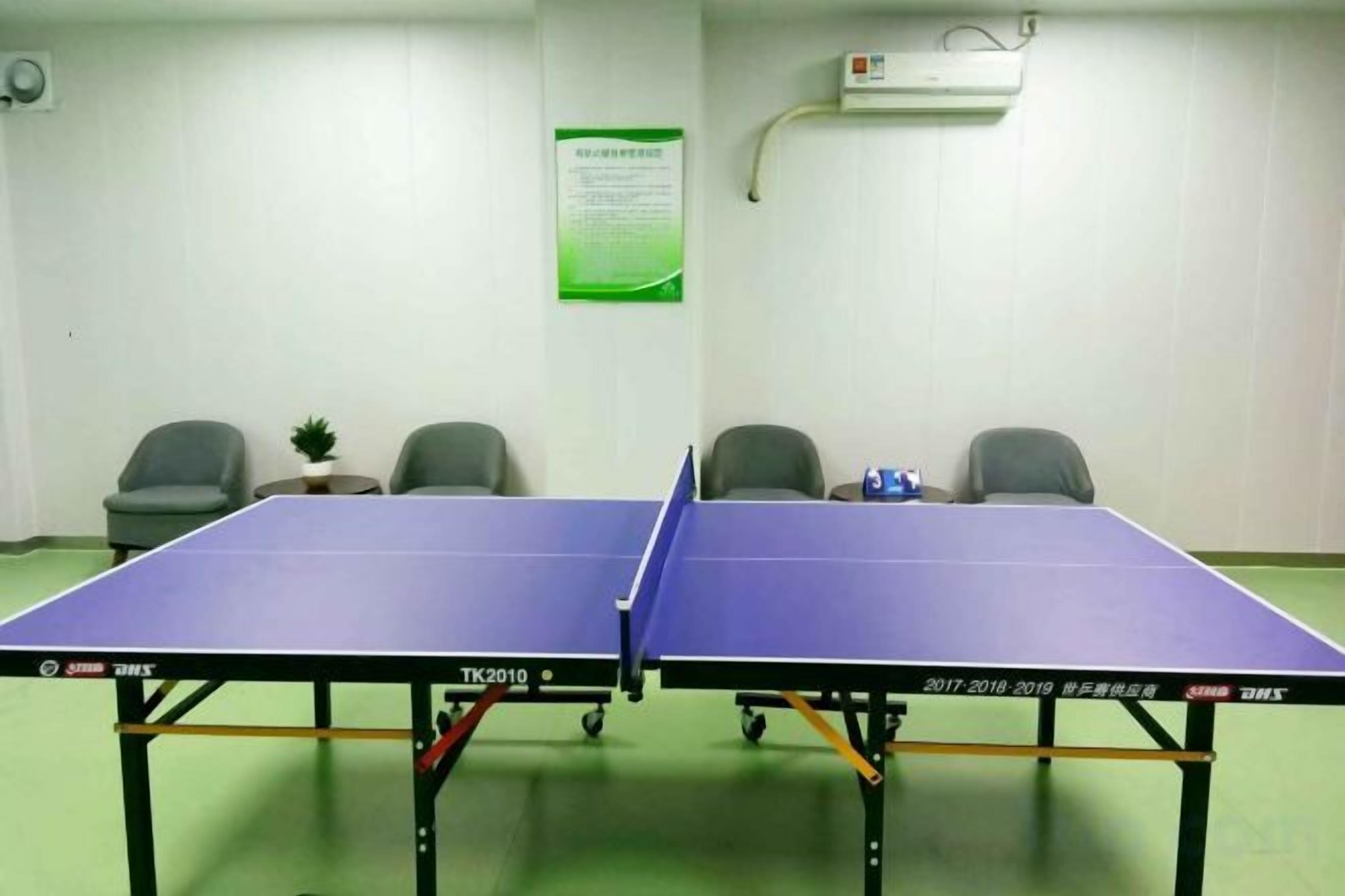 A room with two ping pong tables, a green floor, and four office chairs.