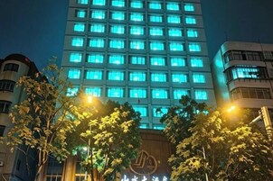 Un edificio de varios pisos con ventanas iluminadas de noche, rodeado de árboles y farolas.