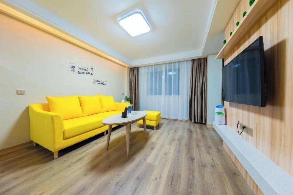 Un salon lumineux avec un canapé jaune, une petite table ronde et un téléviseur fixé au mur.