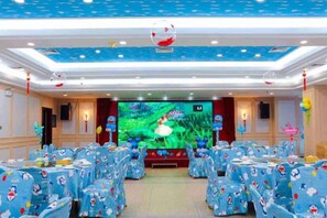 A banquet hall with round tables covered in blue tablecloths, a stage with a large screen, and a ceiling with a blue pattern.