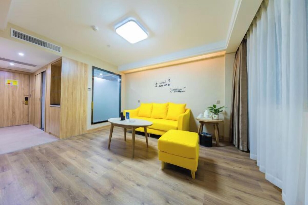 A modern hotel room with a wooden floor, a yellow sofa, a small round table, and a potted plant.