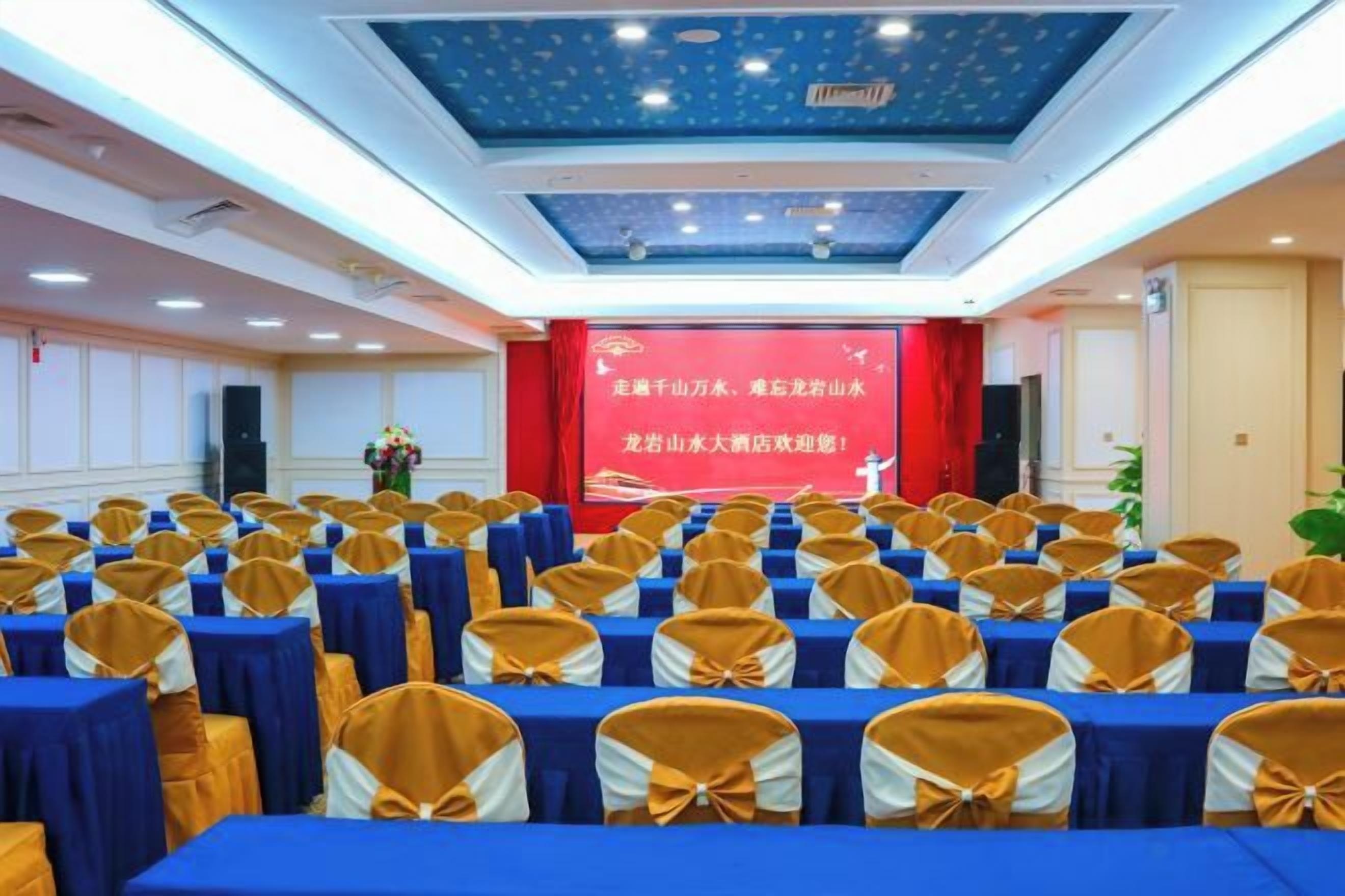 Un salón de conferencias con filas de mesas y sillas, un escenario con un telón de fondo rojo e iluminación empotrada en el techo.