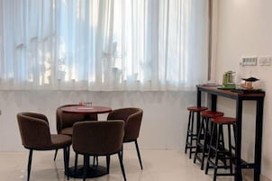 A dining area with a round table and four chairs, a bar counter with stools, and a window with sheer curtains.