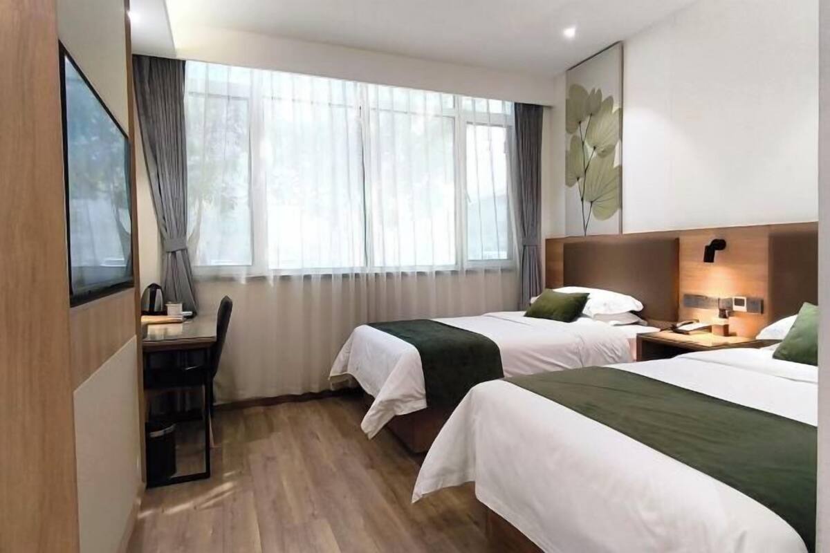 A hotel room with two beds, a desk, a chair, a window with curtains, and a wall-mounted lamp.