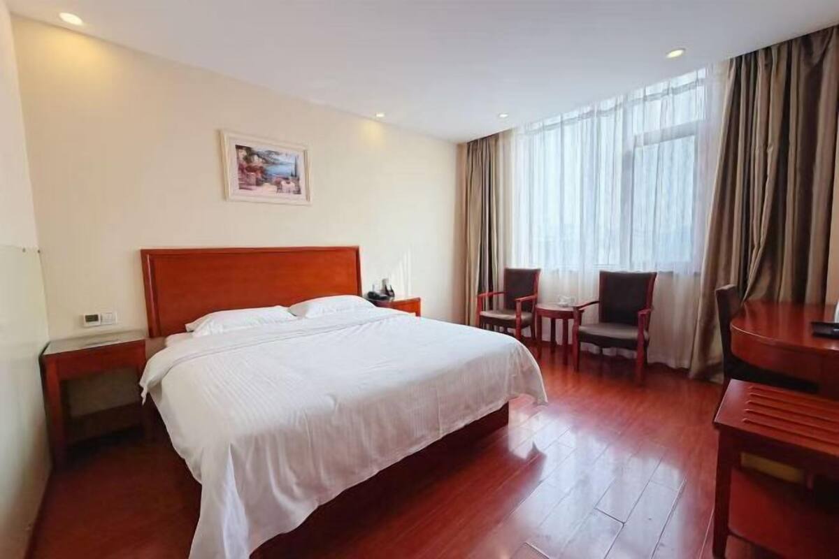 A hotel room with a large bed, a desk, two chairs, a nightstand, and a window with curtains.