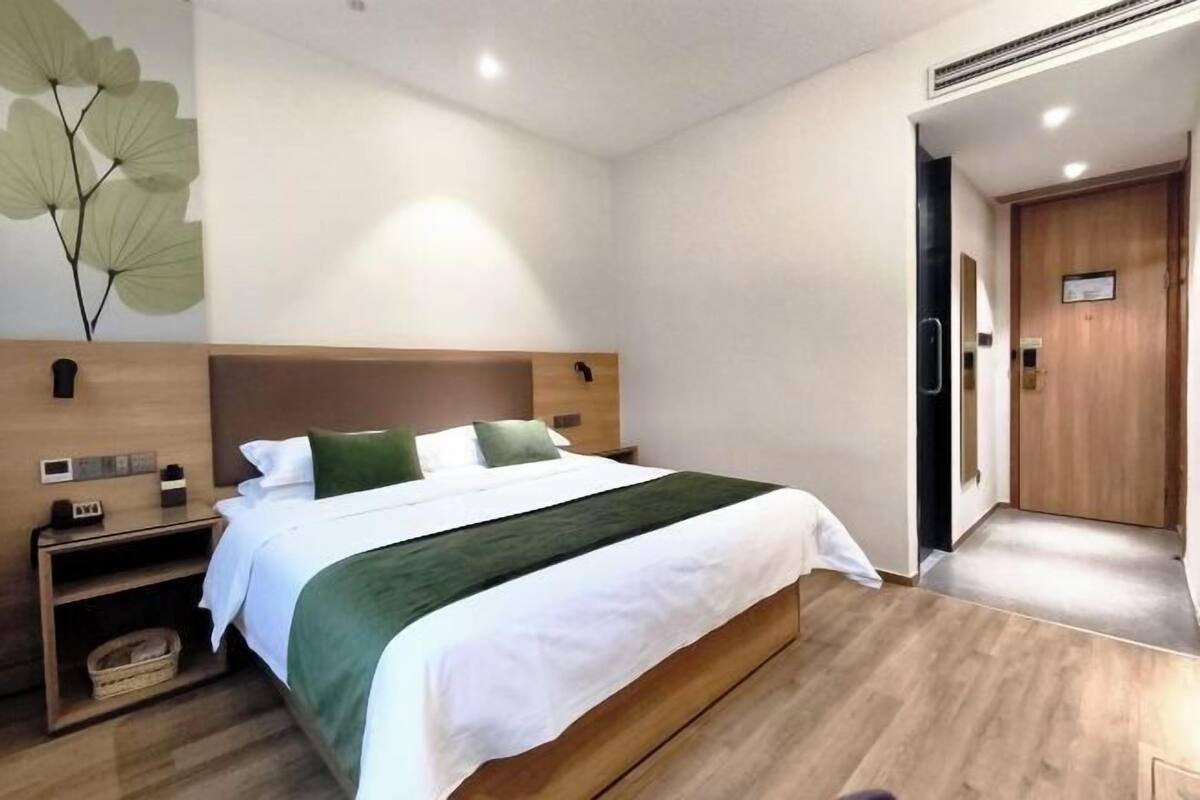 A hotel room with a wooden bed, a nightstand, a wall-mounted clock, and a door to the right.