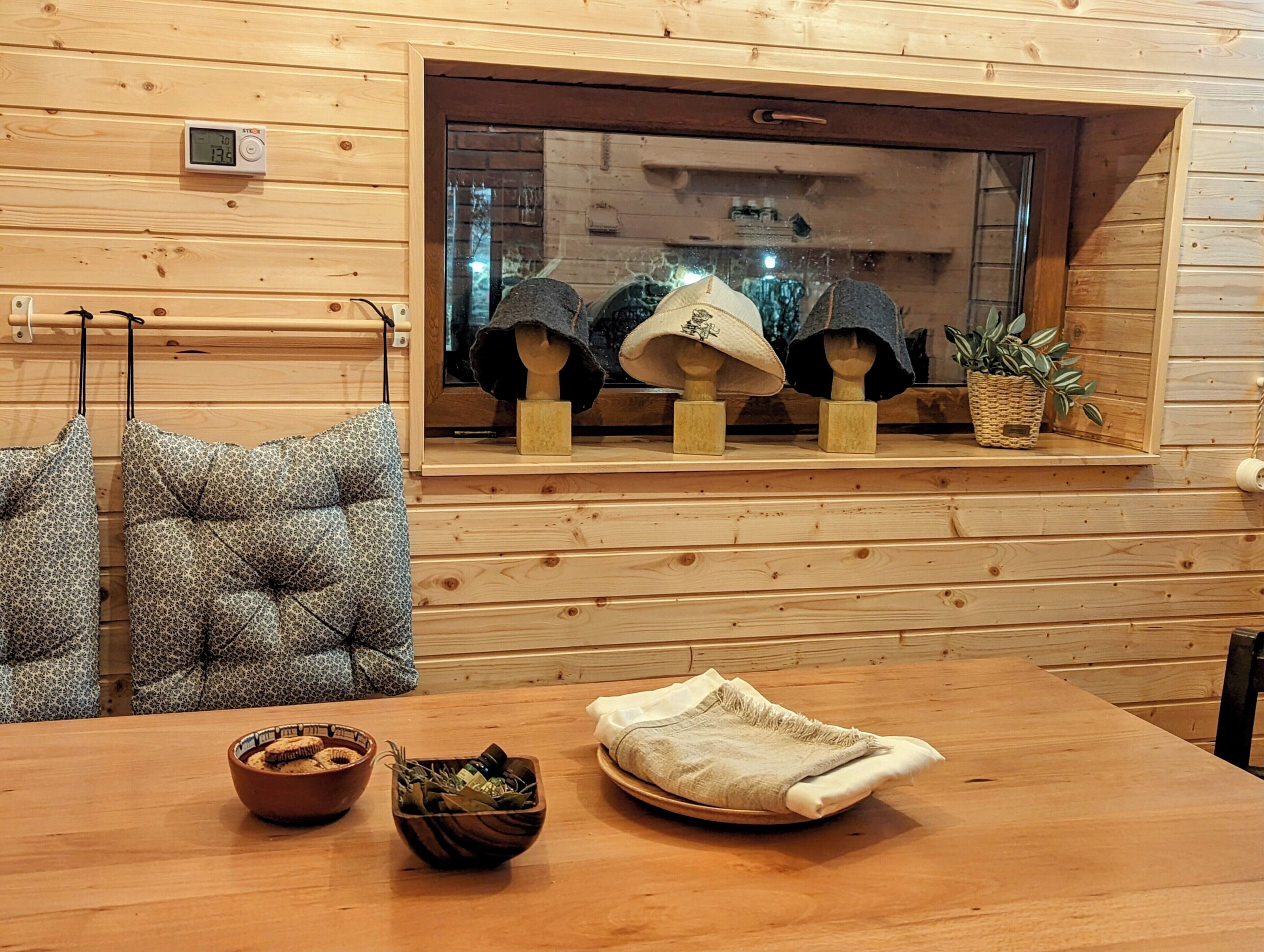 Un interior de madera con una ventana que muestra sombreros y una planta. Hay dos sillas con cojines estampados, una mesa con cuencos y un paño doblado.