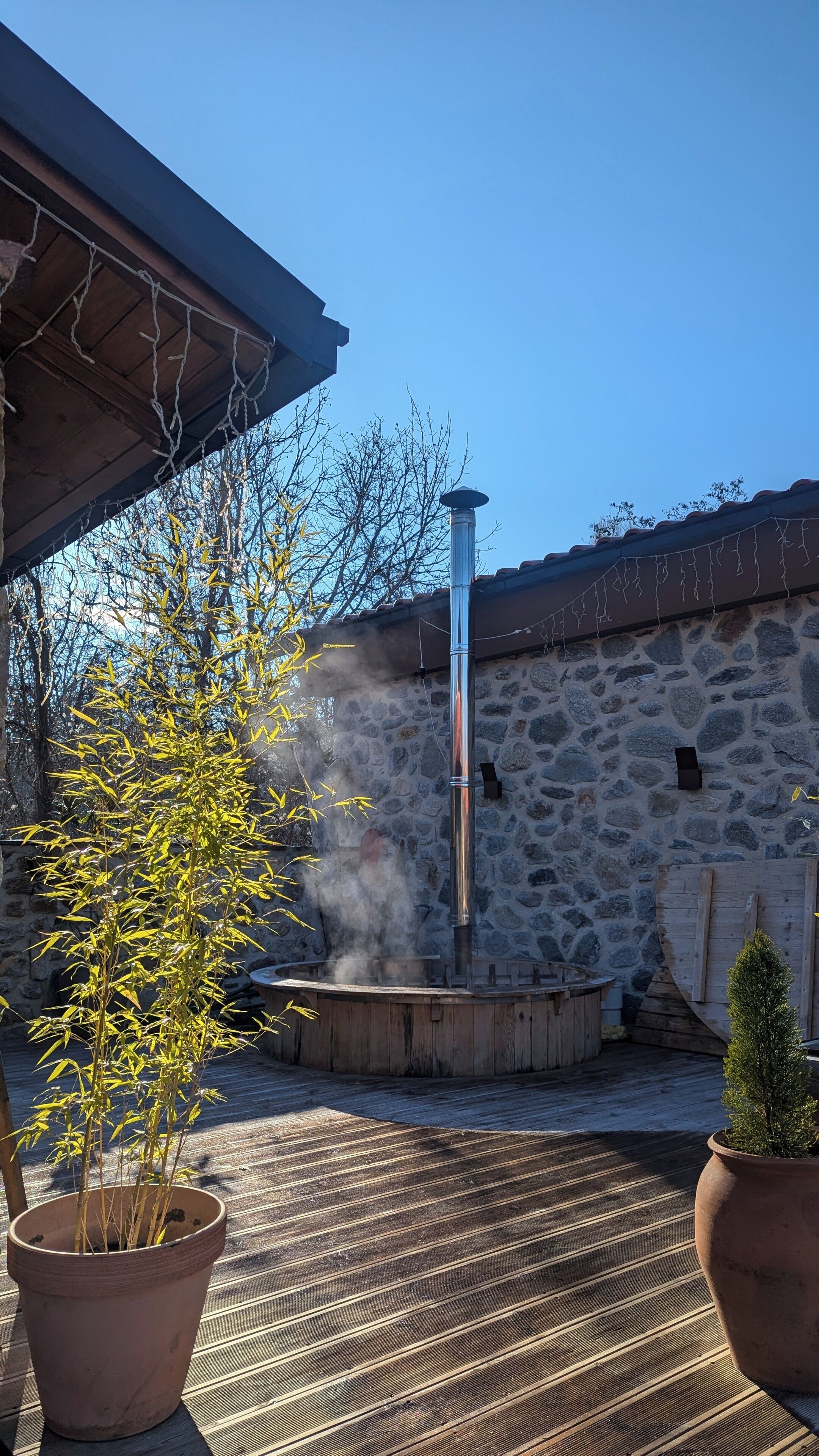 A stone building with a chimney emitting smoke, surrounded by potted plants and a wooden deck.