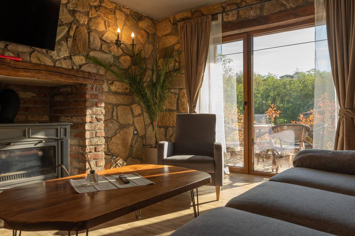 Ein Wohnzimmer mit einer Steinwand, einem Kamin, einer bequemen Couch und einem großen Fenster mit Blick auf Bäume.