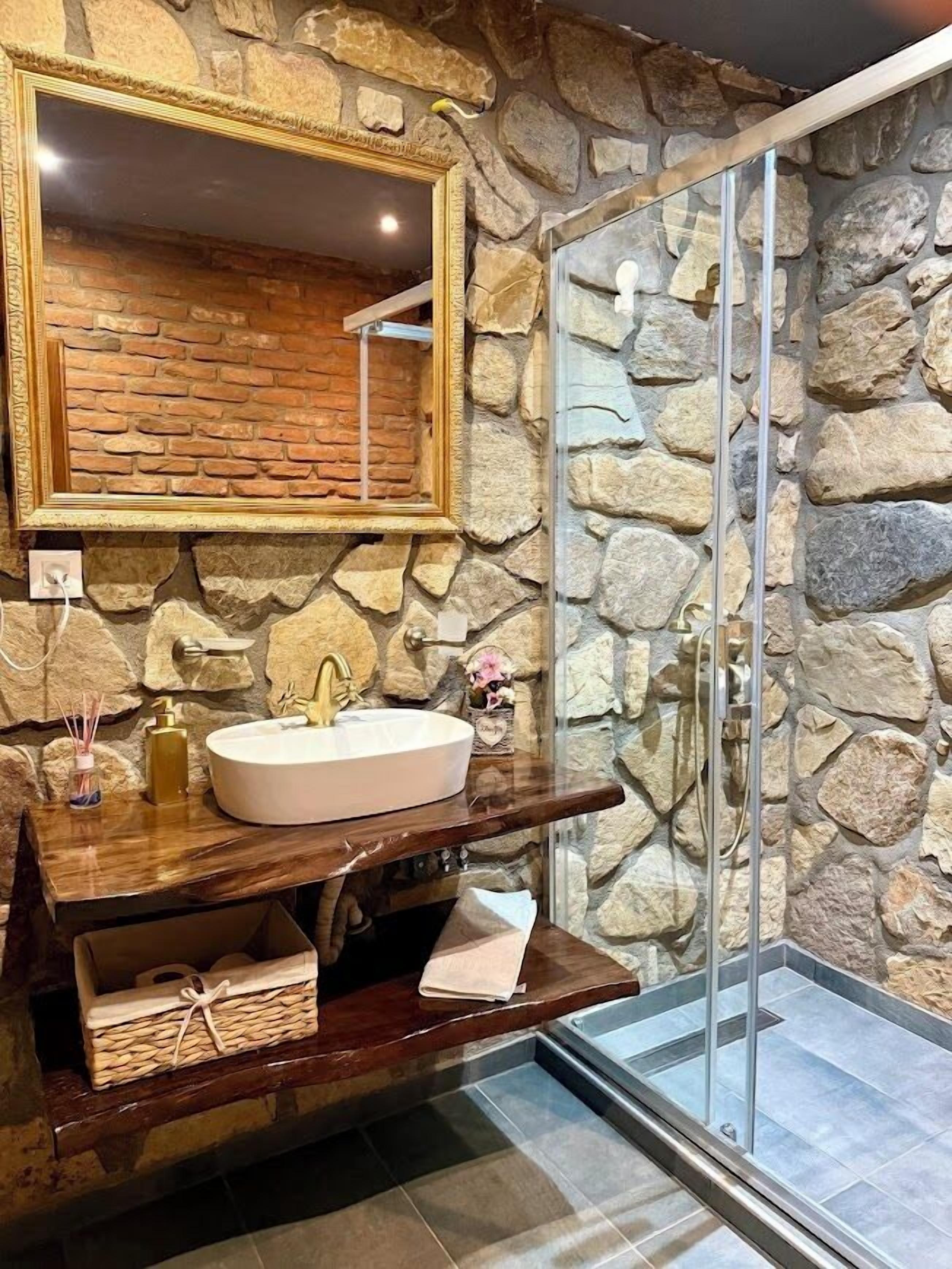 A bathroom with a stone wall, a large mirror, a sink with a gold faucet, and a glass shower enclosure.