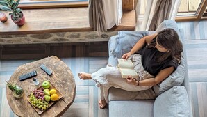 Eine Person liest ein Buch auf einem grauen Sofa in einem Raum mit Holzboden, einem runden Holztisch mit Früchten und einer Fernbedienung, und Blick auf Gartenmöbel aus Holz.