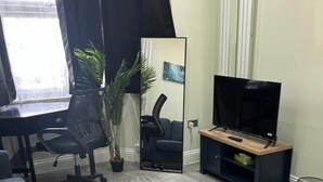 A living room with a TV on a stand, a desk with a chair, a potted plant, and a large mirror. - Tooting Stays (London)