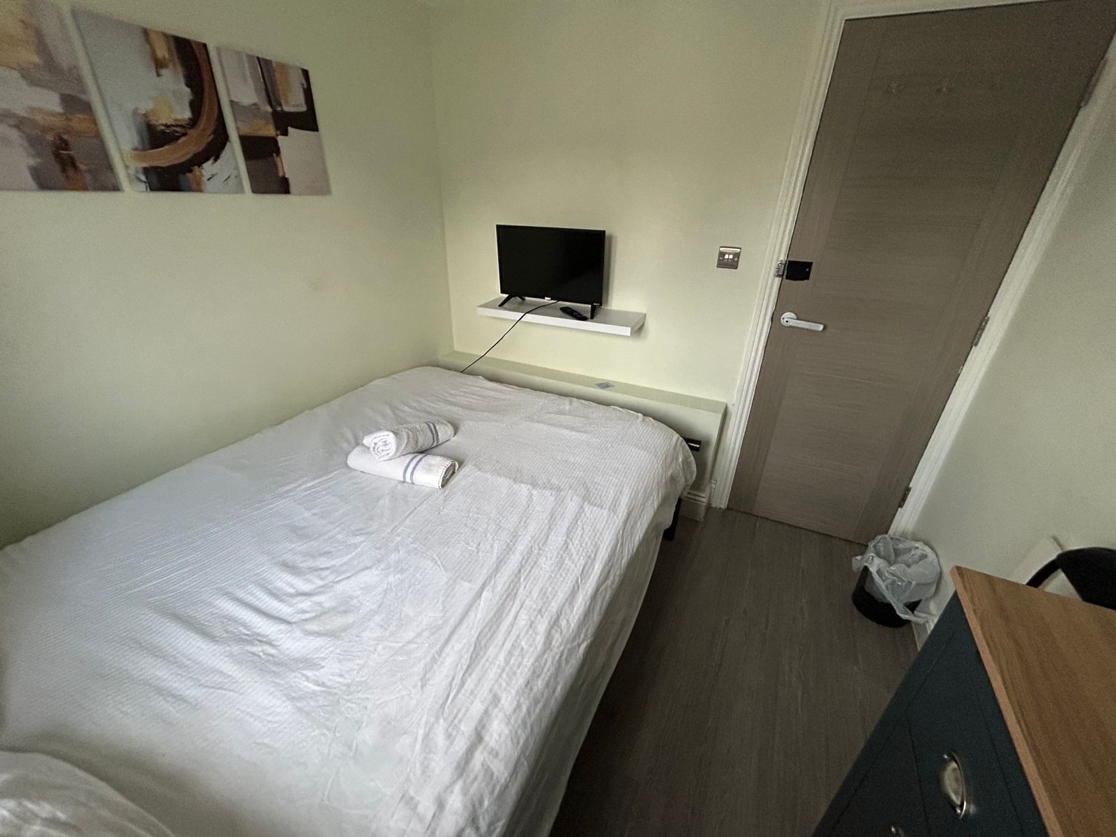 A small, neatly made bed with white linens and two rolled towels. A flat-screen TV is mounted on the wall above the bed.