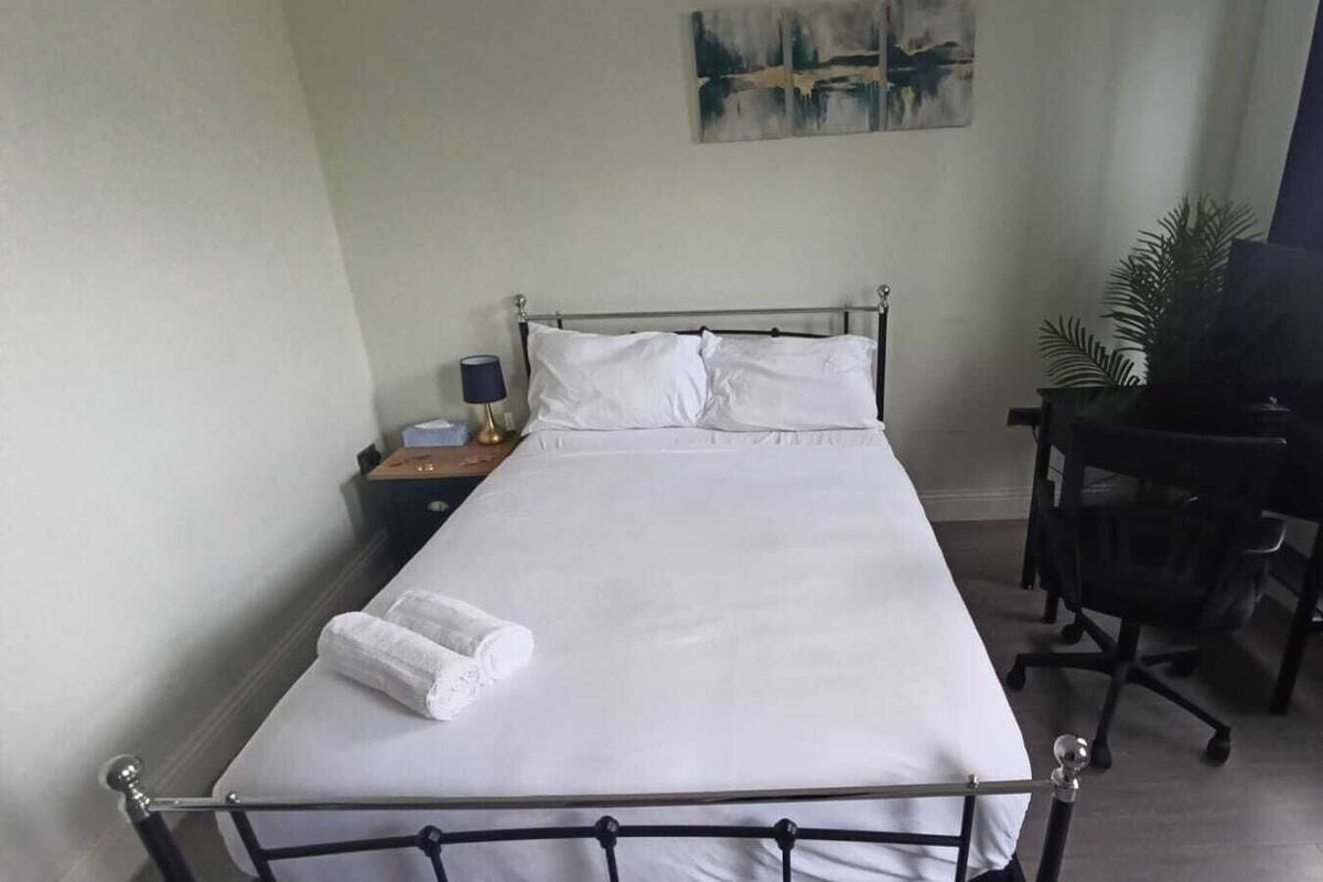 A neatly made bed with white linens, a bedside table with a lamp, a desk with a chair, and a painting on the wall.