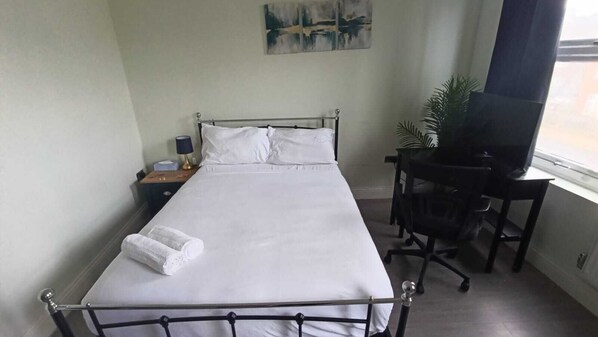 Un lit bien fait, avec du linge de lit blanc, une table de chevet avec une lampe, un bureau avec une chaise et un tableau accroché au mur.
