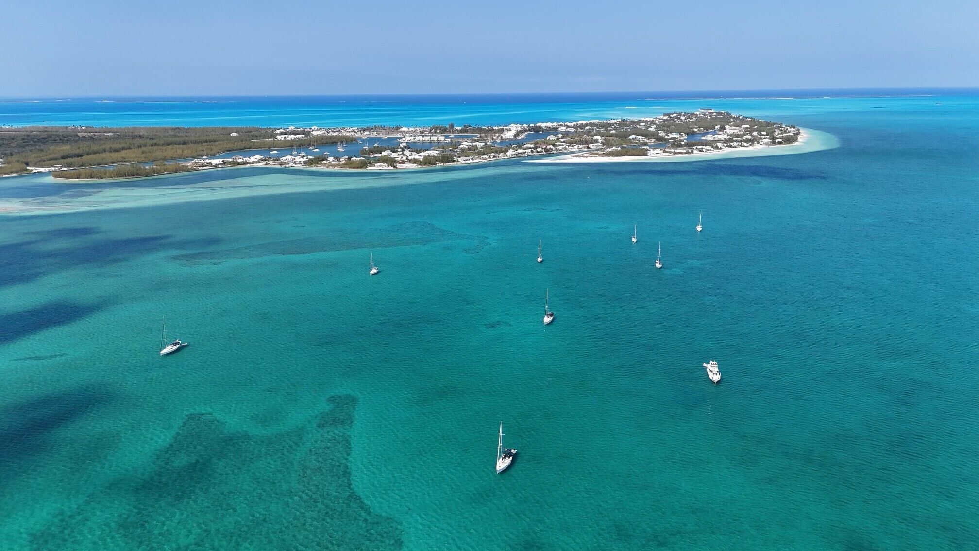 Eine Luftaufnahme einer Küstenlandschaft mit klarem türkisblauem Wasser, mehreren Segelbooten und einer kleinen Insel mit Gebäuden.