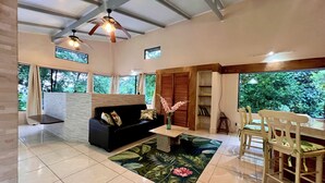 A spacious living area with a black sofa, a dining table with chairs, a ceiling fan, and large windows offering a view of greenery. - Zen Zaru Beautiful Hidden Hotel (Quepos)