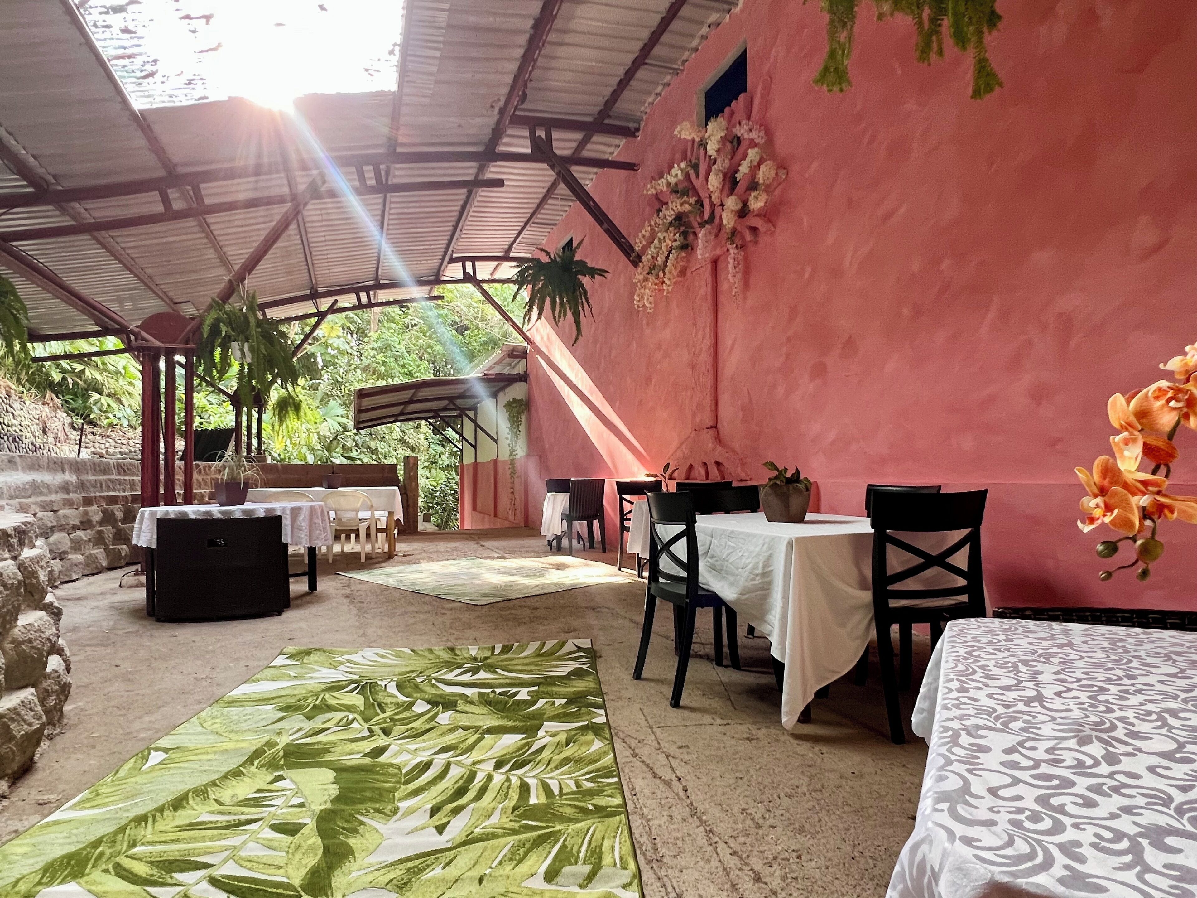 A covered outdoor seating area with tables and chairs, surrounded by stone walls and greenery.