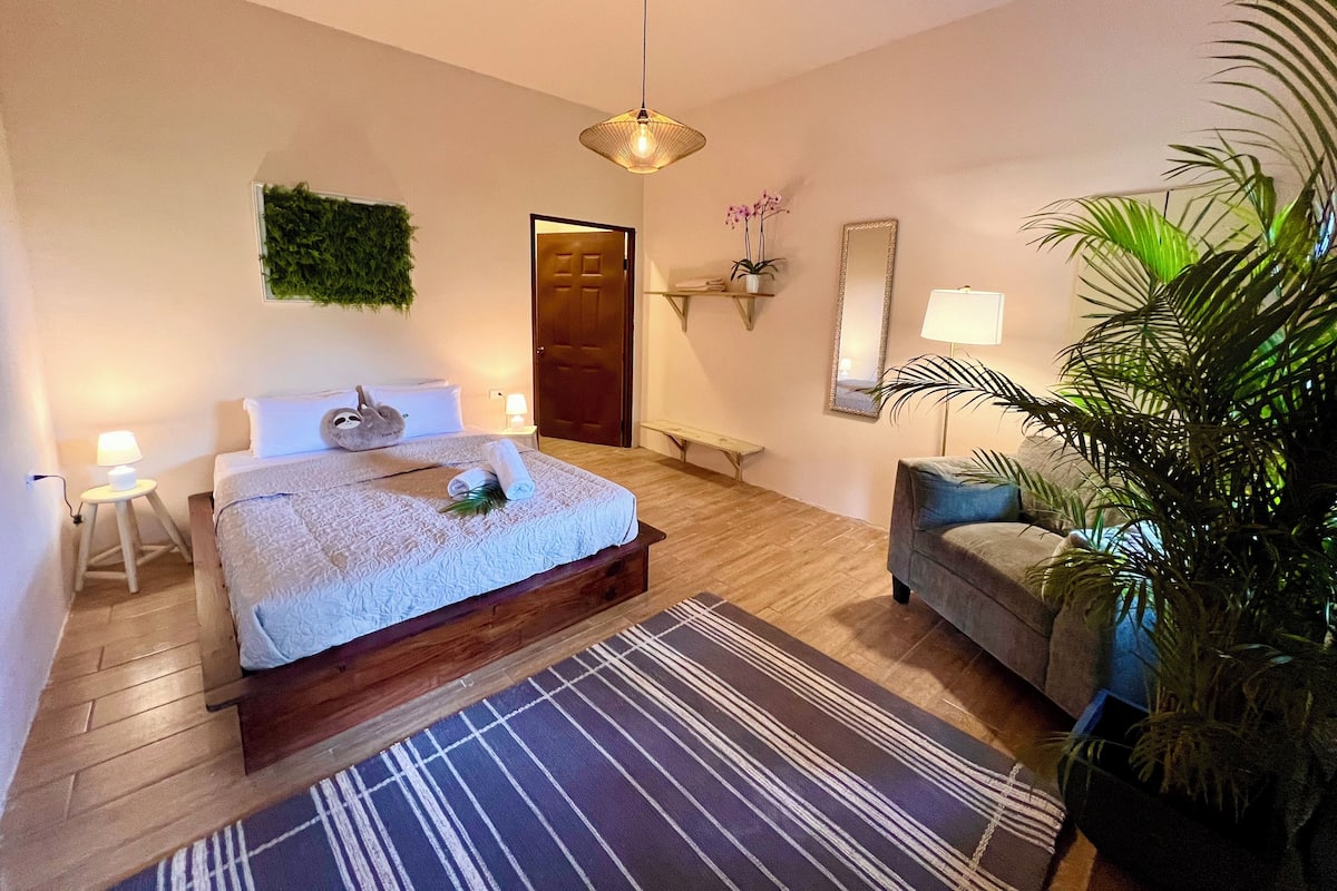 A bedroom with a bed, bedside tables, a bench, a sofa, a mirror, and a potted plant.