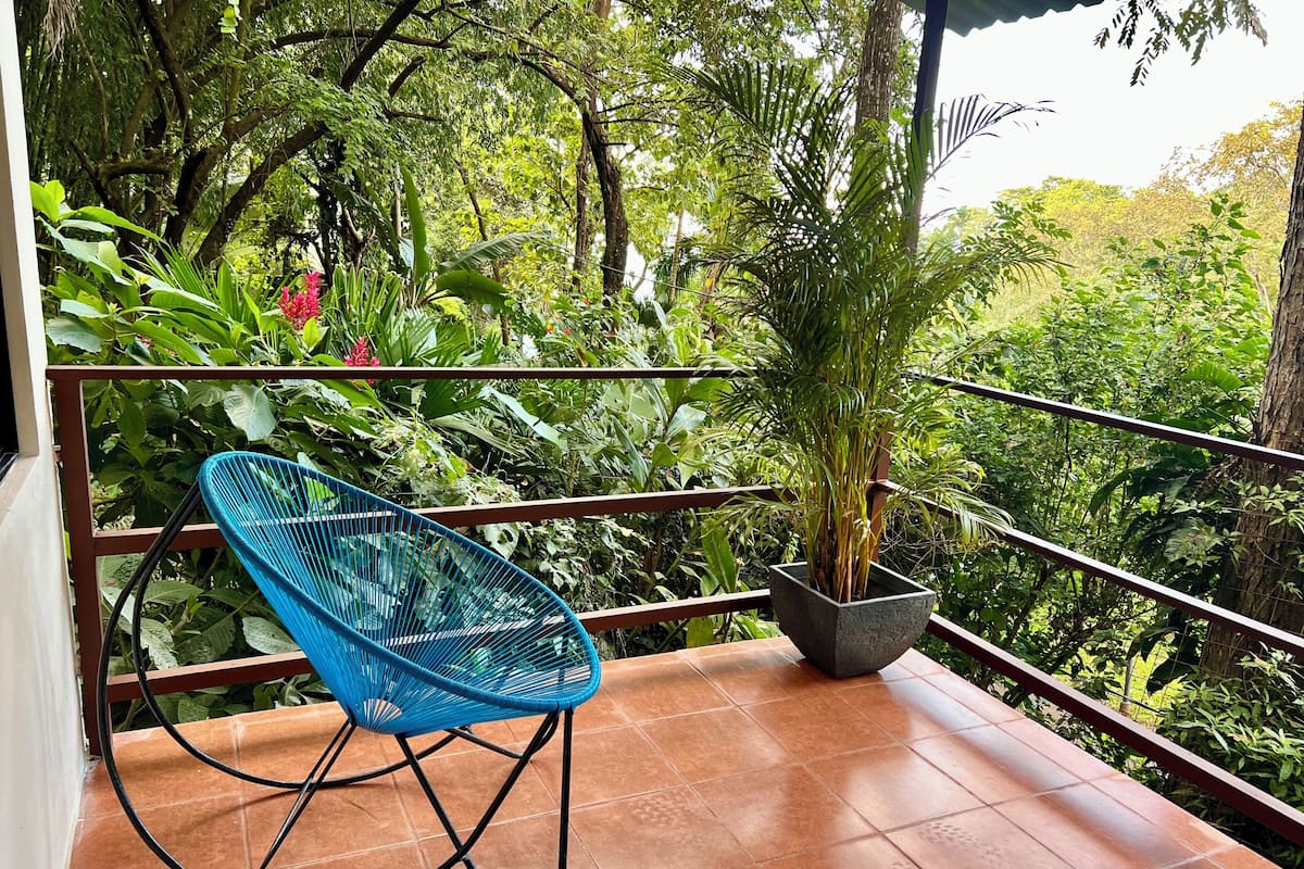 A blue chair on a tiled balcony with a potted plant and lush greenery in the background.