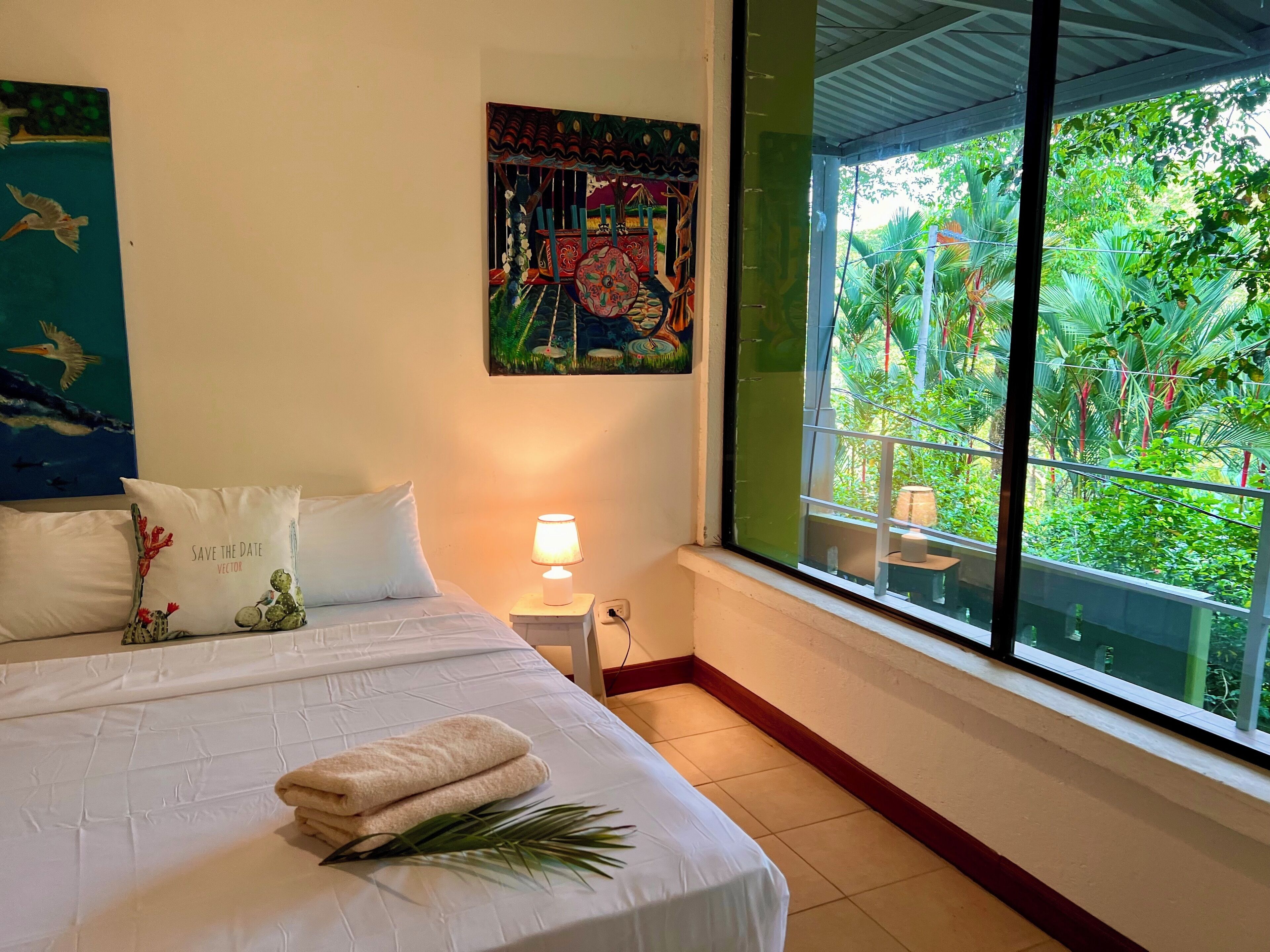 Une chambre d’hôtel avec un lit, des oreillers, une table de chevet avec une lampe, un tableau au mur et une fenêtre donnant sur un paysage verdoyant.