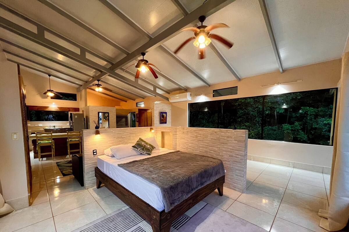 A modern bedroom with a large bed, a ceiling fan, and a view of the outdoors through a large window.