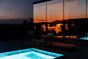 Ett modernt utomhusområde med sittplatser, en pool, ett rum med glasväggar och en säng, samt utsikt över landskapet.