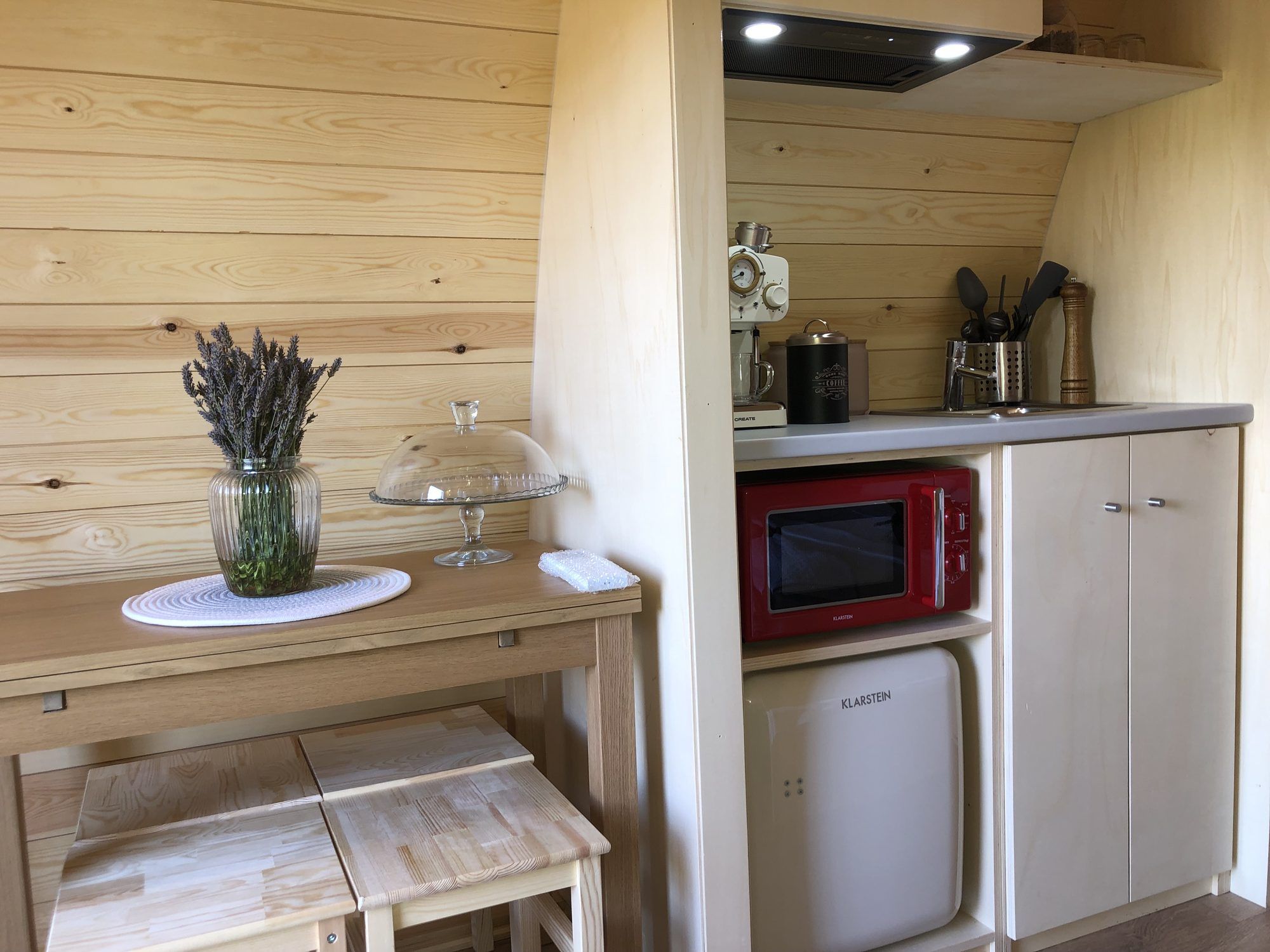 A compact kitchenette with a red microwave, a small refrigerator, and a wooden table with a vase of lavender.