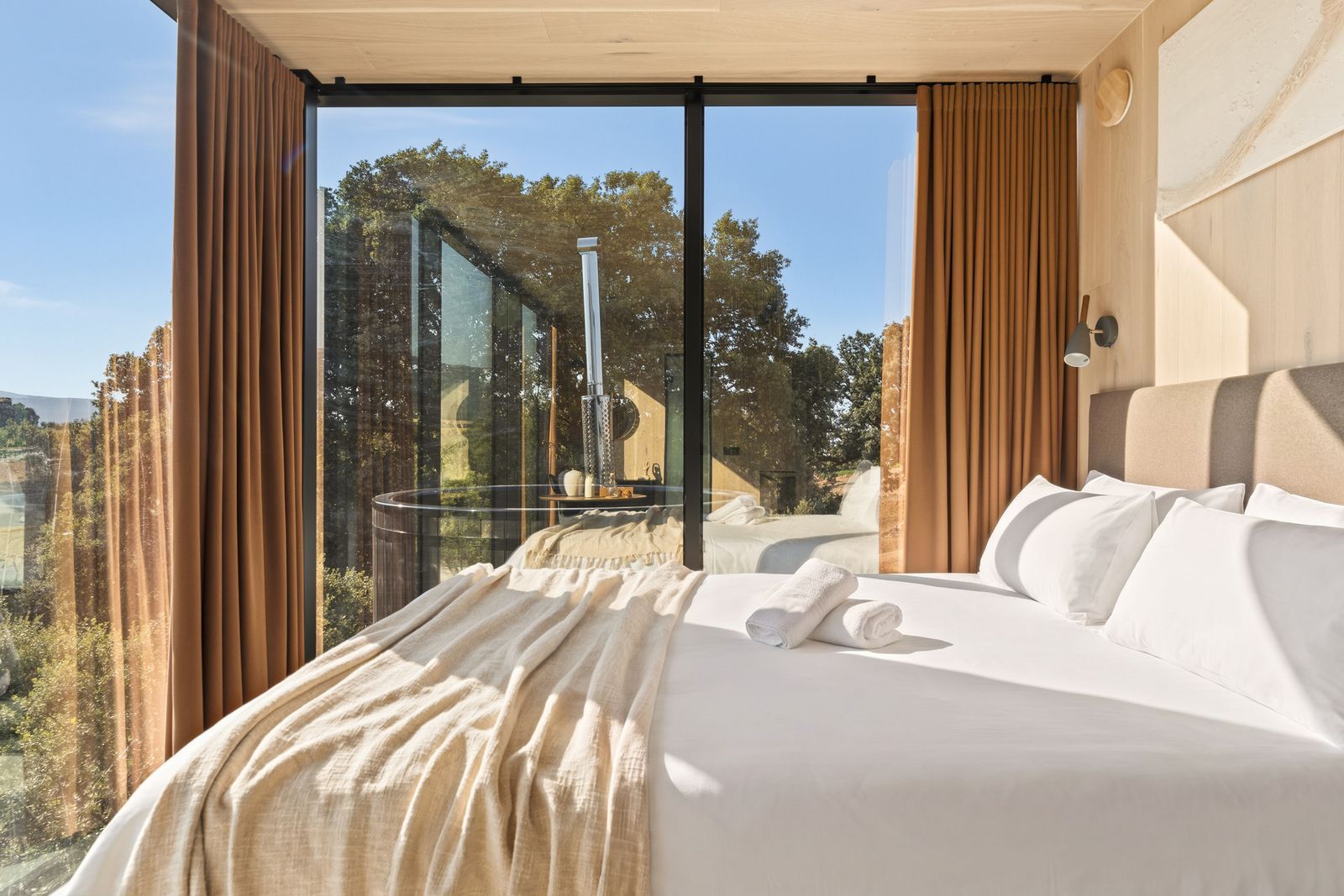 A hotel room with a large bed, a glass wall, and a view of trees and a building.