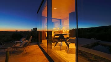 Uma área de estar moderna ao ar livre, com parede de vidro, piscina e vista para o pôr do sol.