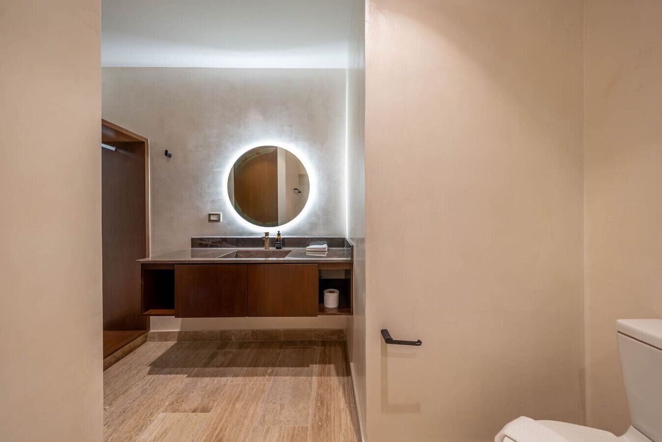 A modern bathroom with a wooden vanity, a round mirror with integrated lighting, and a toilet.