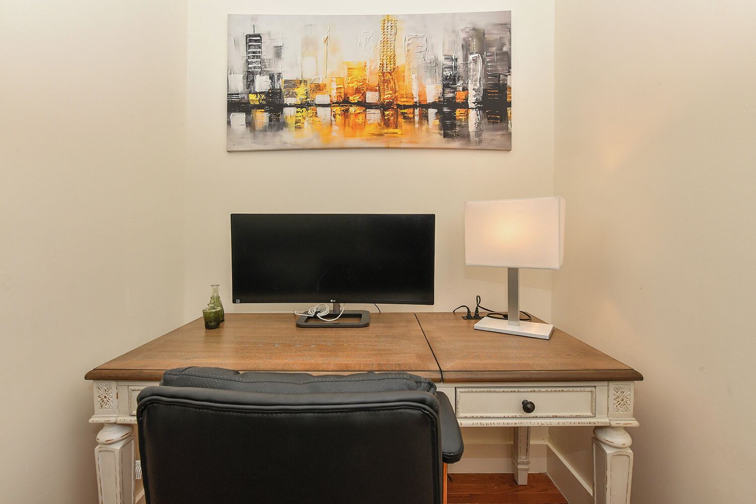A room with a wooden desk, a black chair, a flat-screen TV, a lamp, and a cityscape painting on the wall.