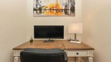 A room with a wooden desk, a black chair, a flat-screen TV, a lamp, and a cityscape painting on the wall.