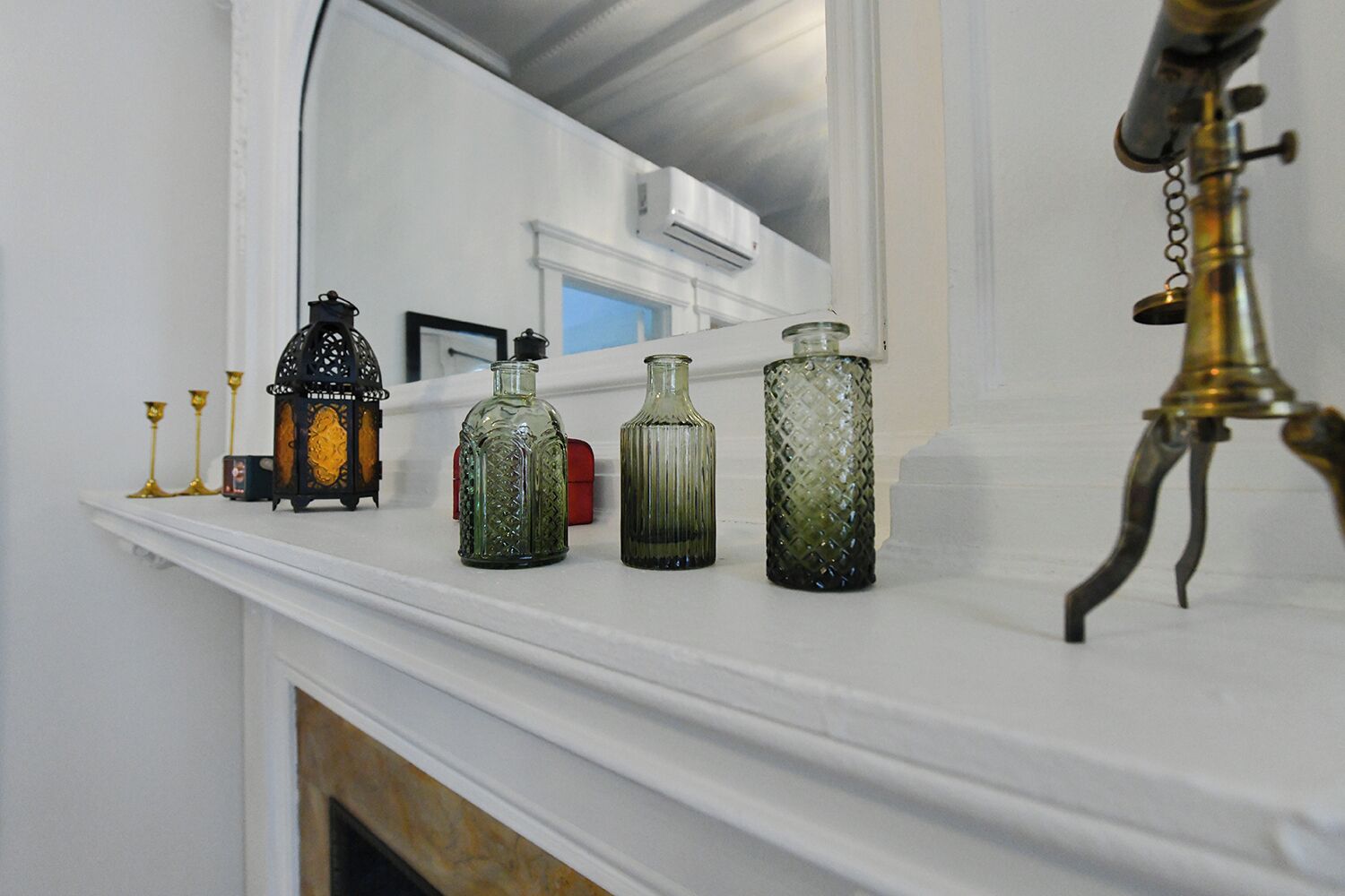Een witte schoorsteenmantel met drie glazen flessen en een messing lamp.