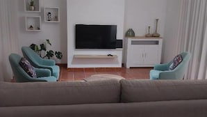 Een woonkamer met een grijze bank, twee turquoise fauteuils, een flatscreen televisie op een witte standaard, en een kast met decoratieve objecten.
