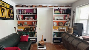 A cozy living room with a black leather sofa, a purple armchair, and a wooden coffee table. There are bookshelves filled with books and decorative items, and a television on a wooden stand. - Charming 1-bedroom private suite. Easy walk to beach and boardwalk. (Virginia Beach)