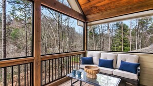 Une terrasse en bois avec une table en verre, un canapé blanc avec des coussins bleus et une vue sur une zone boisée.