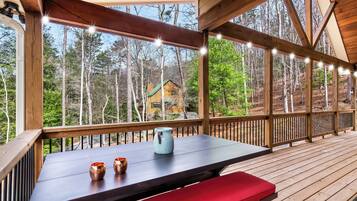 A wooden deck with a table and benches, overlooking a forested area with a cabin.