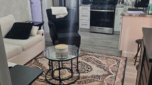 A compact living space with a sofa, a chair, a glass table, a patterned rug, a kitchen area with appliances, and a door leading to another room. - Charming studio in superb Fort Myers that’s conveniently located (Fort Myers)