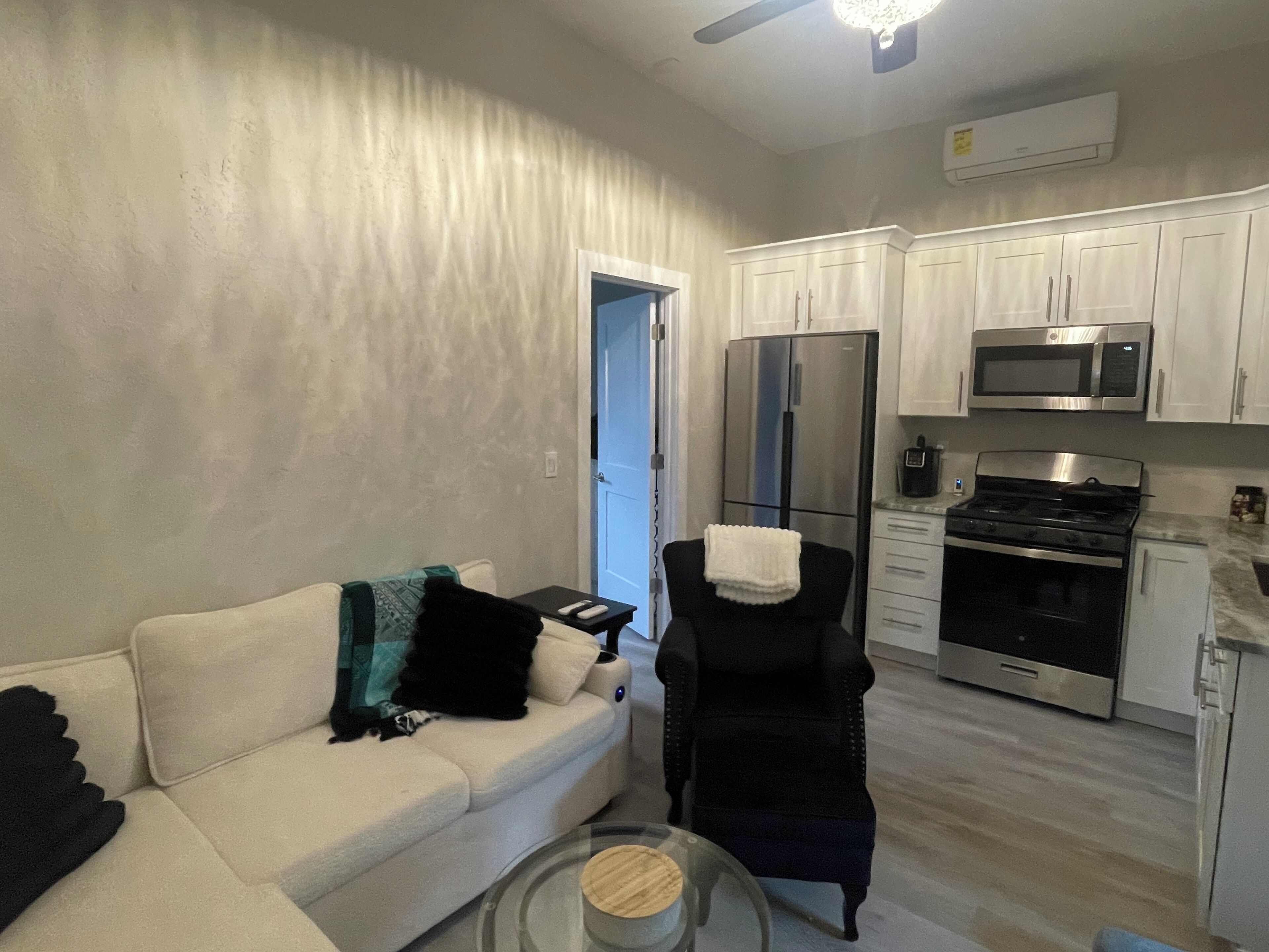 A modern living room with a white sofa, a black armchair, and a glass coffee table. A kitchen with white cabinets, a refrigerator, and a microwave is visible in the background.