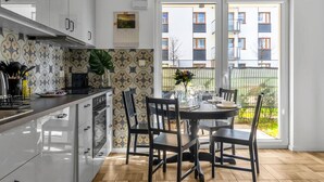 Eine moderne Küche mit weißen Schränken, ein Essbereich mit rundem Tisch und Stühlen sowie Blick auf ein Wohngebäude durch eine Glastür.